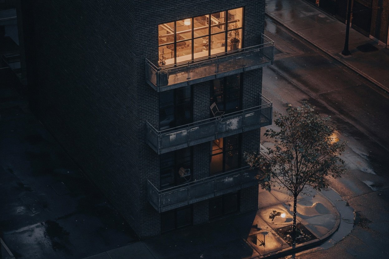 A narrow dark-brick building at night, with a brightly lit top floor and slim balconies.